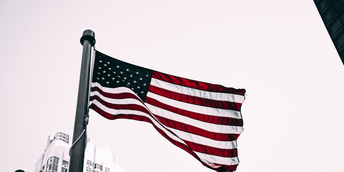 American flag waving on a flagpole with city buildings visible in the background.