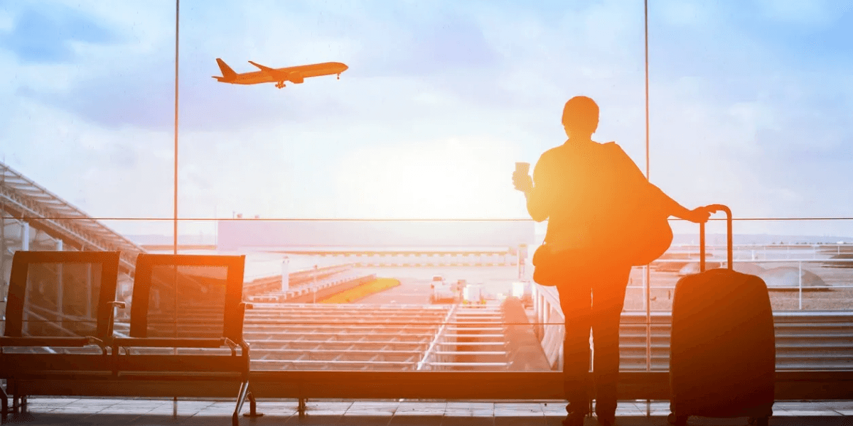 **Traveler with suitcase watches departing airplane through airport terminal window at sunrise.**