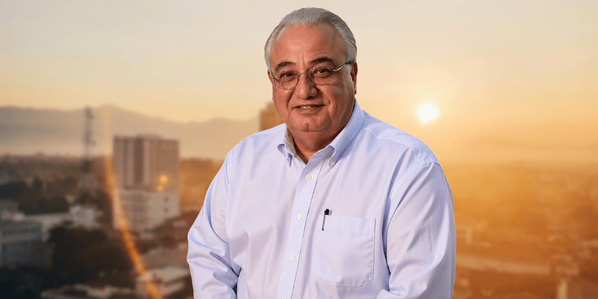 Portrait of Kenneth Carnesi, Sr. smiling in a light blue shirt against a blurred city skyline at sunset.