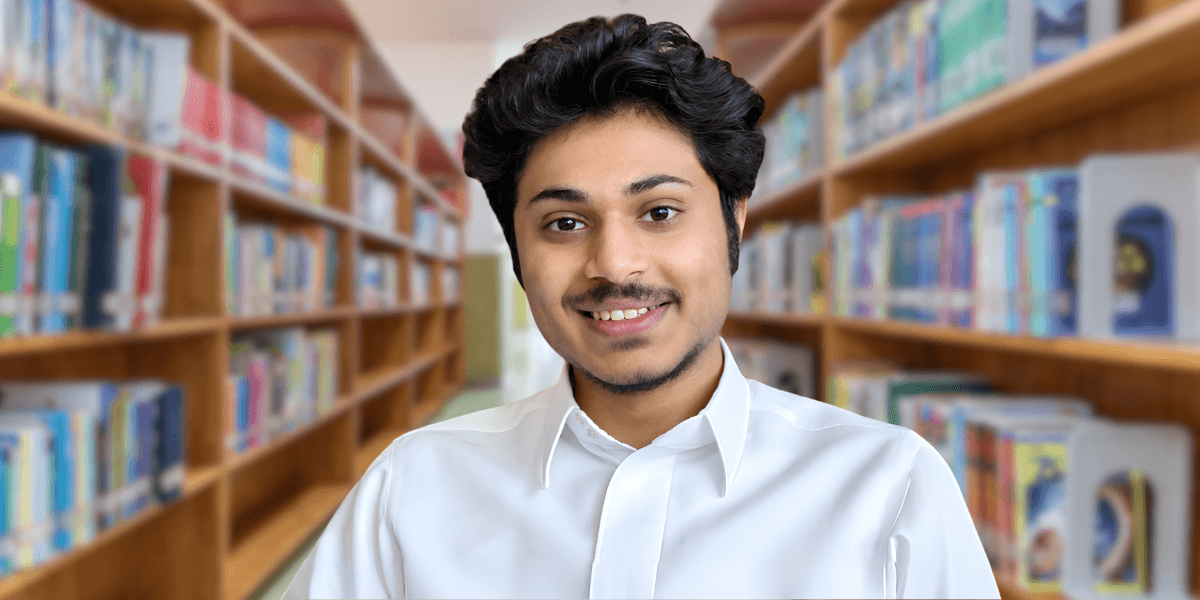 Young man in a white shirt smiling in a library with bookshelves behind.