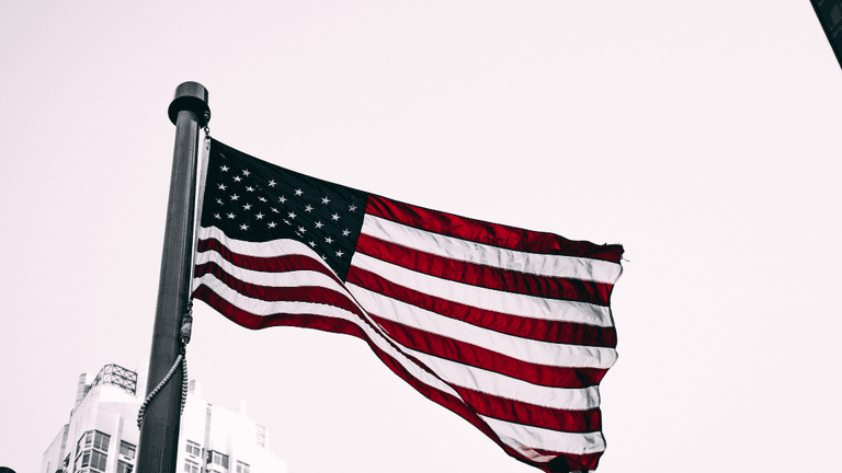 American flag waving on a flagpole with city buildings visible in the background.