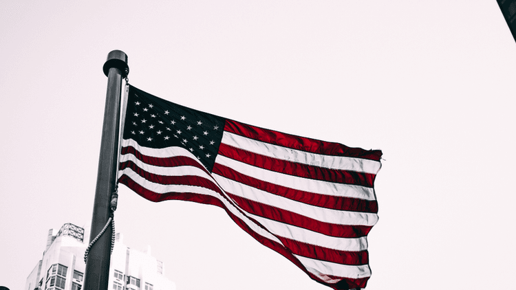 American flag waving on a flagpole with city buildings visible in the background.