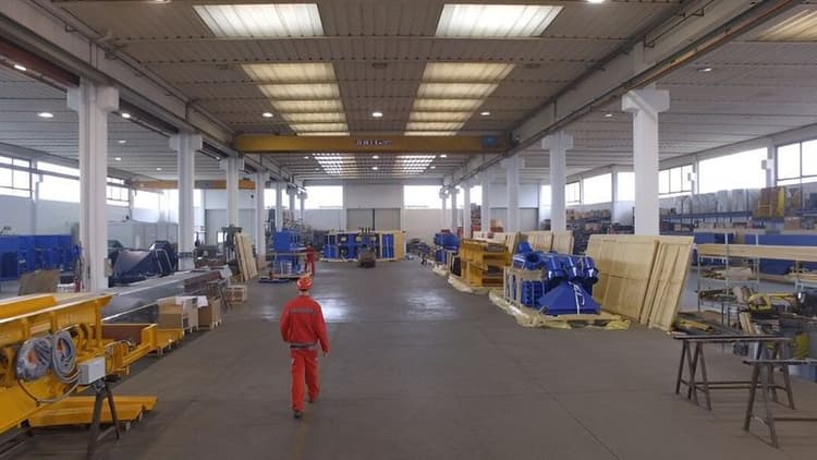 Spacious industrial warehouse with a high ceiling, filled with yellow machinery and equipment. A person in a red uniform walks down the central aisle.