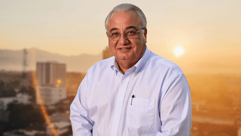 Portrait of Kenneth Carnesi, Sr. smiling in a light blue shirt against a blurred city skyline at sunset.