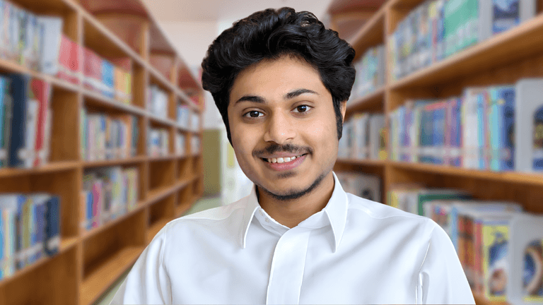 Young man in a white shirt smiling in a library with bookshelves behind.