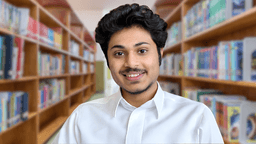 Young man in a white shirt smiling in a library with bookshelves behind.