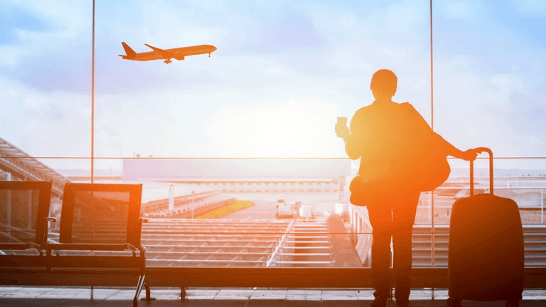 **Traveler with suitcase watches departing airplane through airport terminal window at sunrise.**