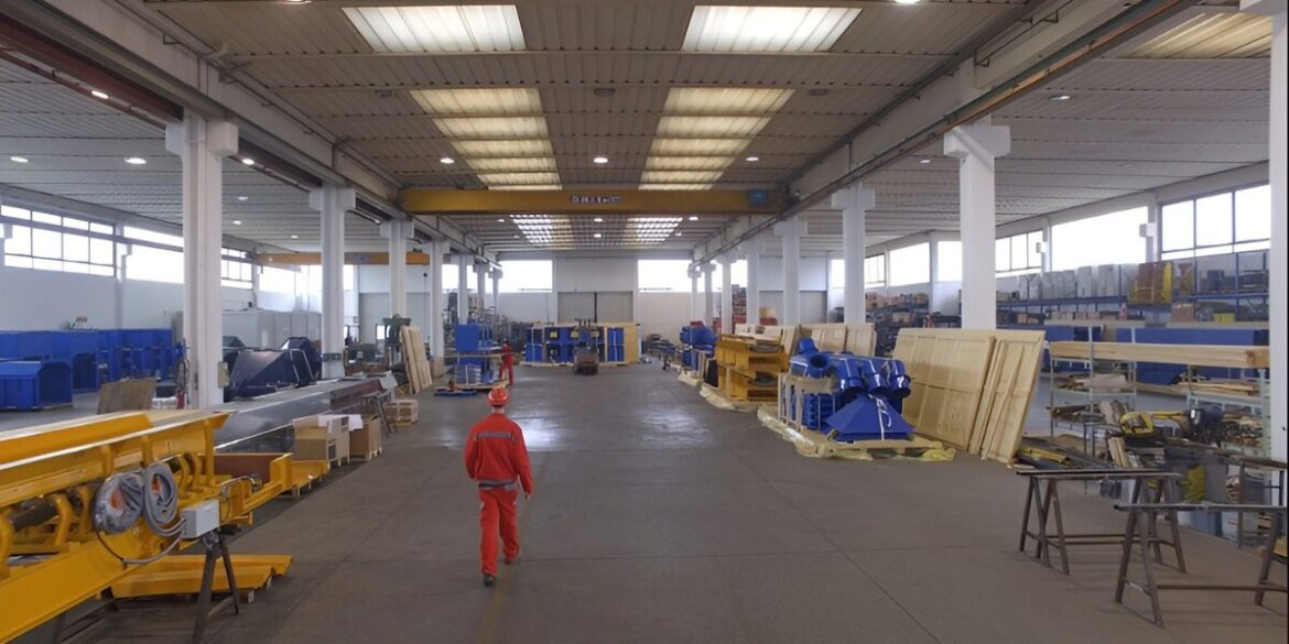 Spacious industrial warehouse with a high ceiling, filled with yellow machinery and equipment. A person in a red uniform walks down the central aisle.