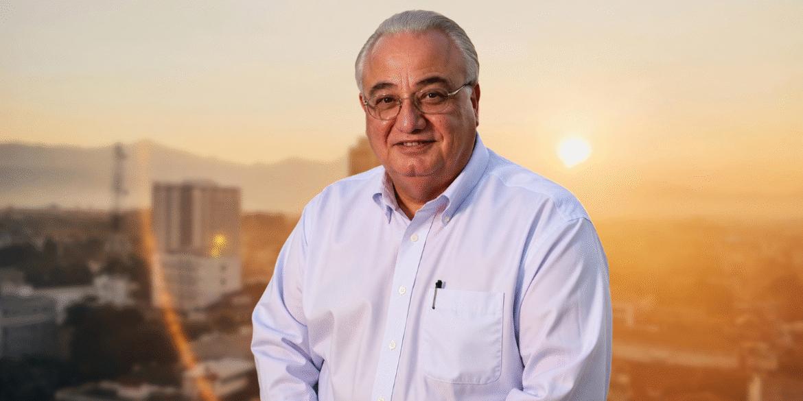 Portrait of Kenneth Carnesi, Sr. smiling in a light blue shirt against a blurred city skyline at sunset.